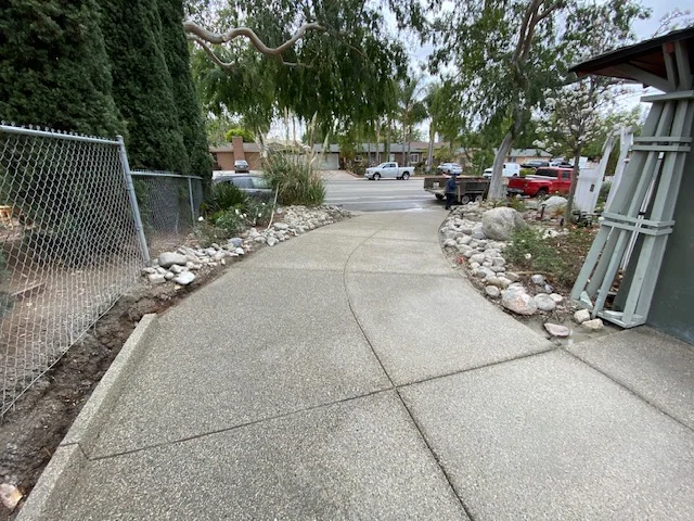 Finished concrete driveway with control joints, rock borders, and chain-link fence integration